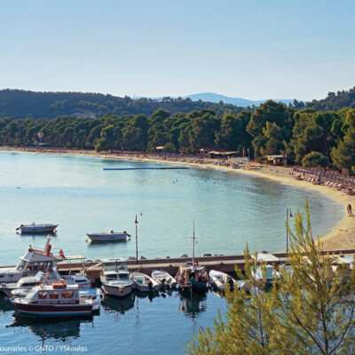 Un ponton avec des bateaux de plaisance, une plage en forme de croissant bordé de pins, le ciel bleu et la mer Egée, à Skiathos, en Grèce