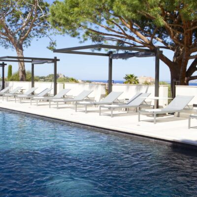 Vue d'une longue piscine bordée de transats, sous l'ombre de pins parasols