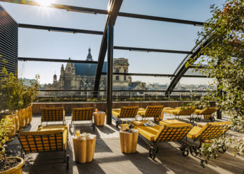 Le rooftop ROOF de l'hôtel Madame Rêve à Paris, avec des chaises longues recouvertes de coussins jaunes, des tables basses en bois avec des jus de fruits et une vue sur les toits de la capitale et l'église Saint-Eustache