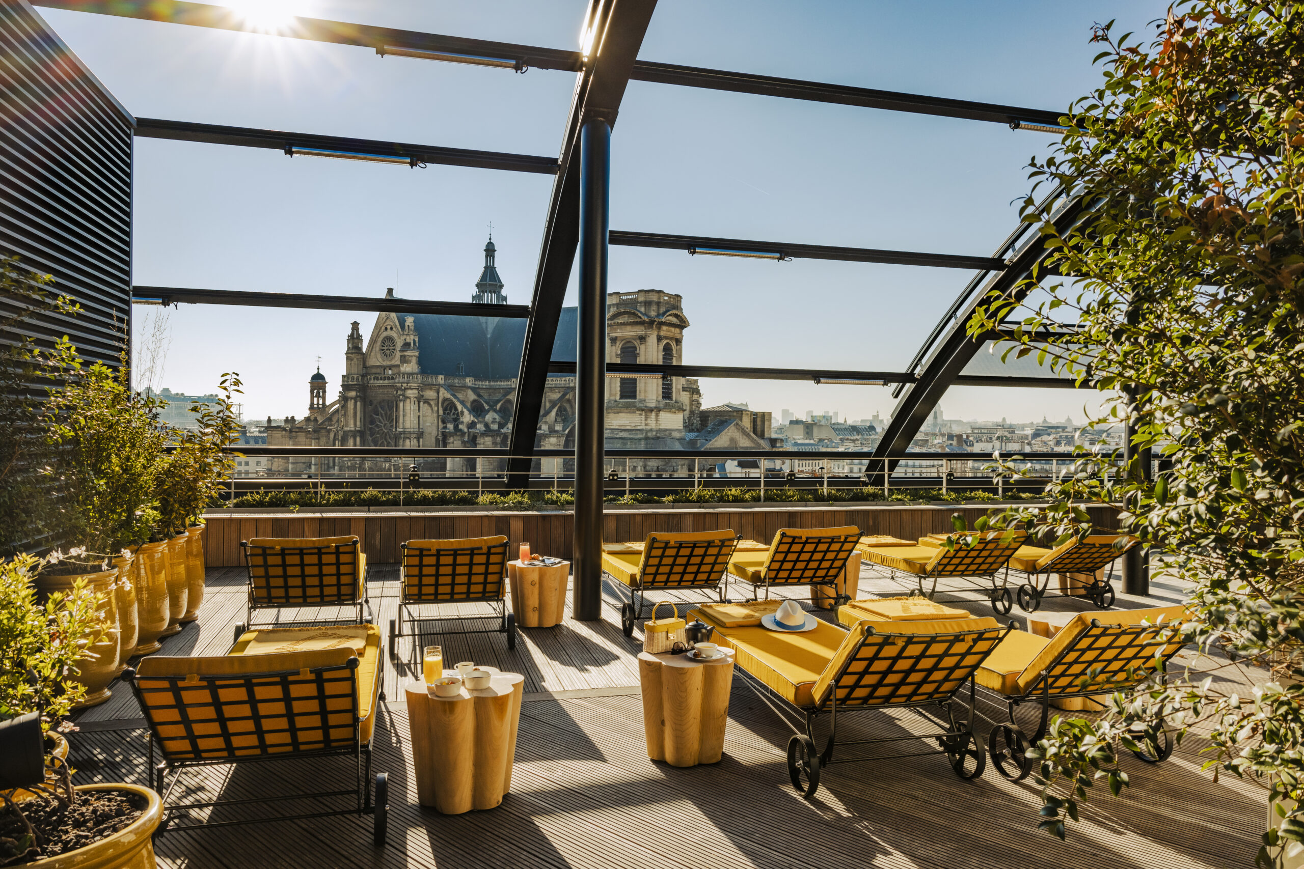 Le rooftop ROOF de l'hôtel Madame Rêve à Paris, avec des chaises longues recouvertes de coussins jaunes, des tables basses en bois avec des jus de fruits et une vue sur les toits de la capitale et l'église Saint-Eustache