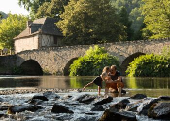 Vue de la rivière qui traverse le village de Le Saillant, avec une vieille maison et un pont en pierre, et deux personnes les pieds dans l'eau, lors d'un séjour dans la région de Brive