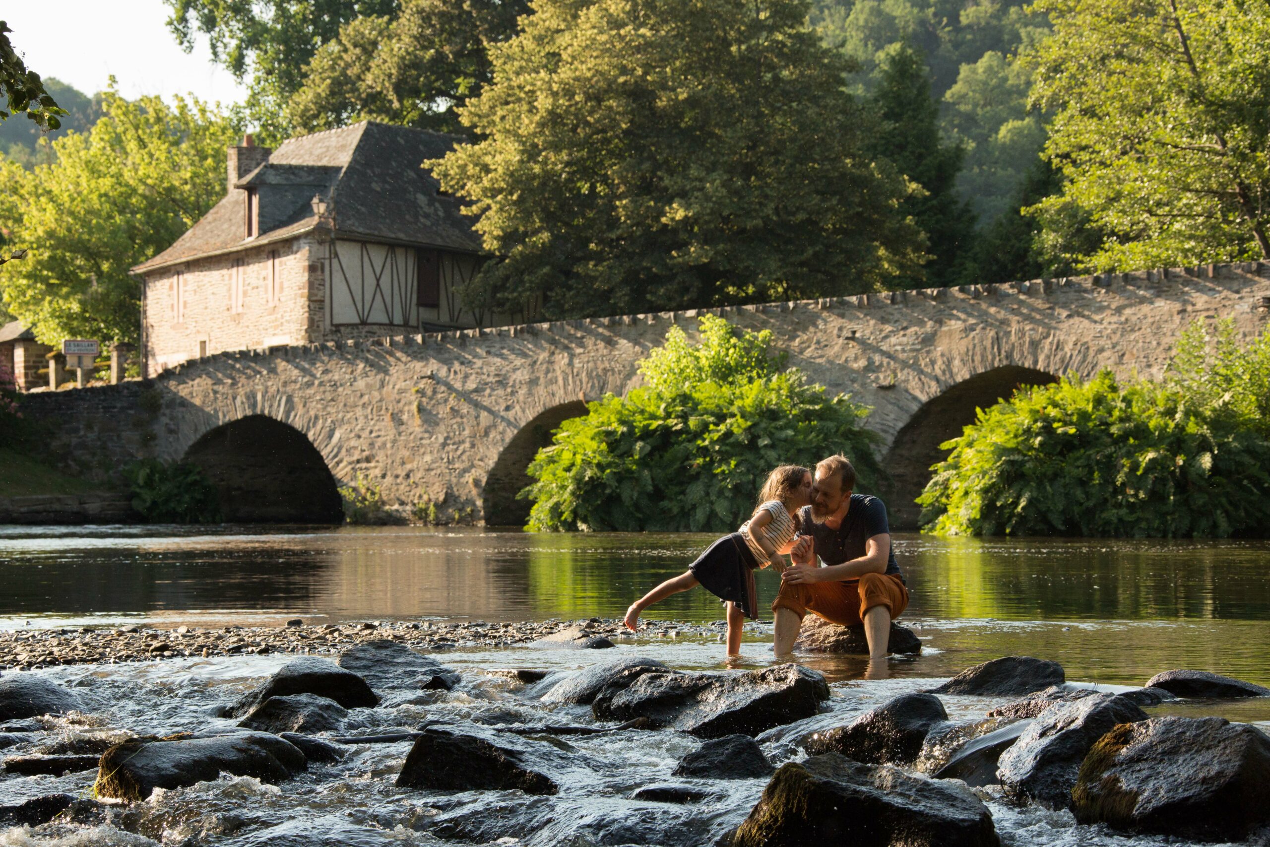 Vue de la rivière qui traverse le village de Le Saillant, avec une vieille maison et un pont en pierre, et deux personnes les pieds dans l'eau, lors d'un séjour dans la région de Brive