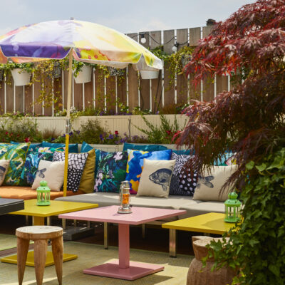 Des tables multicolores, avec des coussins aux motifs de poissons et des parasols fleuris sur le rooftop en bois du Mamma Shelter Paris-East