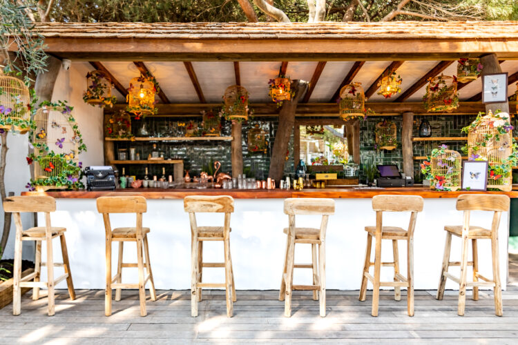 Un bar de plein air avec des chaises hautes en bois, des cocktails, et des cages à oiseaux fleuries, sur la plage privée de Nikki Beach, à St Tropez