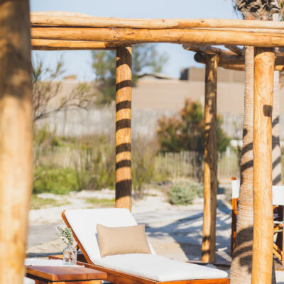 Une chaise longue avec matelas blanc et coussin beige sous une treille en bois, sur la plage privée de Nikki Beach, à St Tropez