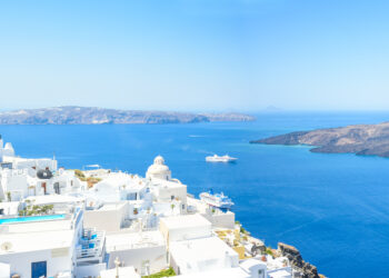 Une vue aérienne d'un village aux maisons et clochers blancs de l'île de Santorin avec en contrebas des îles, la mer Egée et le Lyria, un bateau de la compagnie Ponant qui propose les plus belles croisières estivales