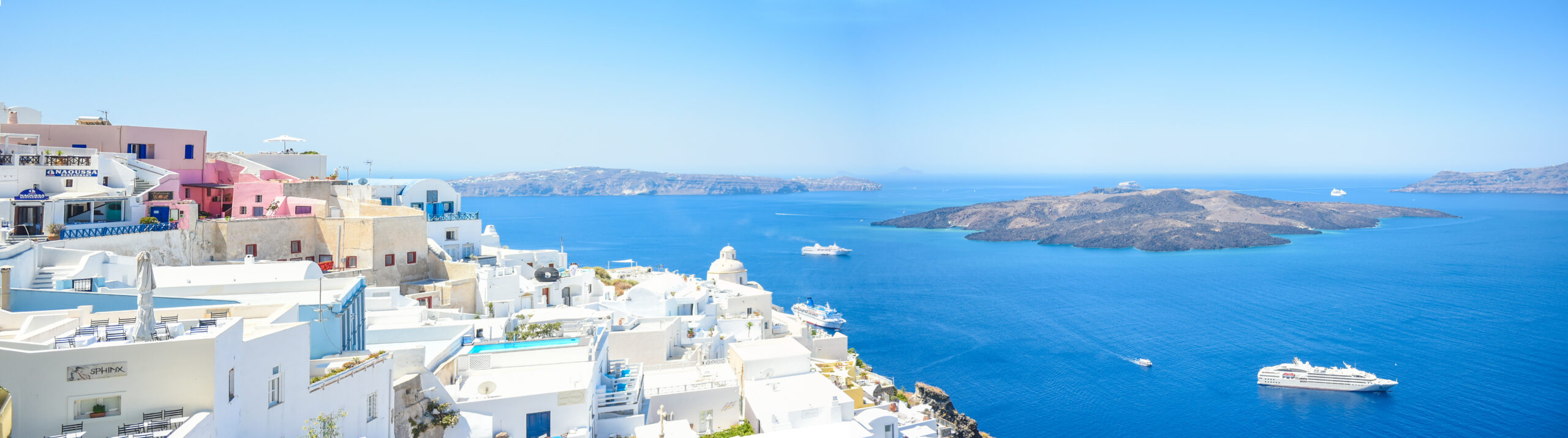 Une vue aérienne d'un village aux maisons et clochers blancs de l'île de Santorin avec en contrebas des îles, la mer Egée et le Lyria, un bateau de la compagnie Ponant qui propose les plus belles croisières estivales