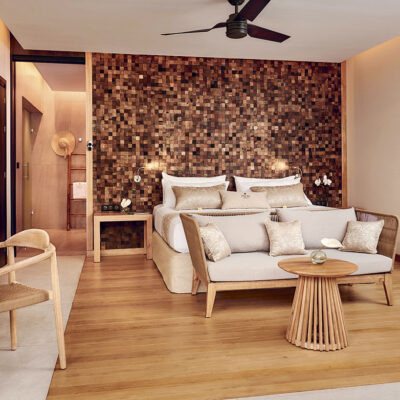 Vue intérieure d'une chambre de l'un des pool-lodges du Palm Hotel & Spa à La Réunion avec du mobilier design de bois clair, un lit, un canapé, un bureau et un murs de mosaïques dorées