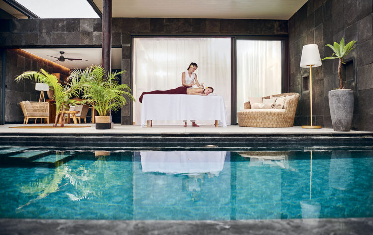 Une longue piscine devant laquelle une femme, couchée sur une table de massage, est en train de se faire masser par une masseuse, près de l'un des pool-lodges du Palm Hotel & Spa à La Réunion