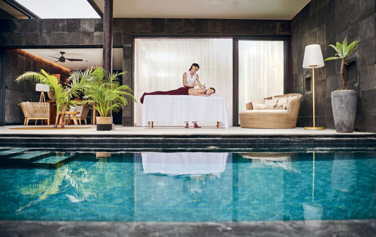 Une longue piscine devant laquelle une femme, couchée sur une table de massage, est en train de se faire masser par une masseuse, près de l'un des pool-lodges du Palm Hotel & Spa à La Réunion