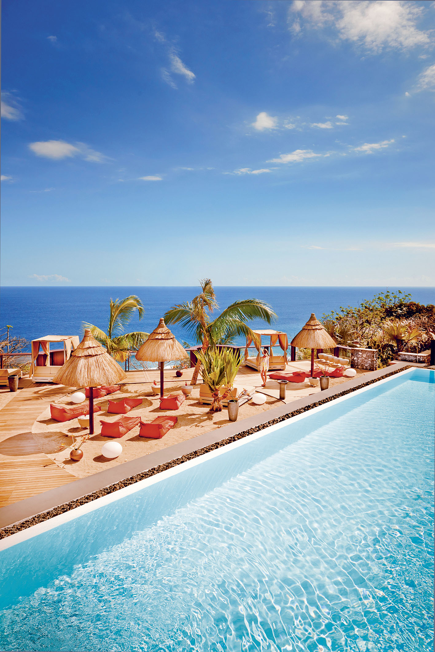 Vue de l'une des piscines du Palm Hotel & Spa à La Réunion, avec des transats, des parasols de feuilles de palmiers et au loin, l'océan Indien