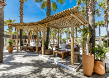 Des canapés, fauteuils et tables basses à l'ombre de treilles en bois et de palmiers, sur la plage privée de Nikki Beach, à St Tropez