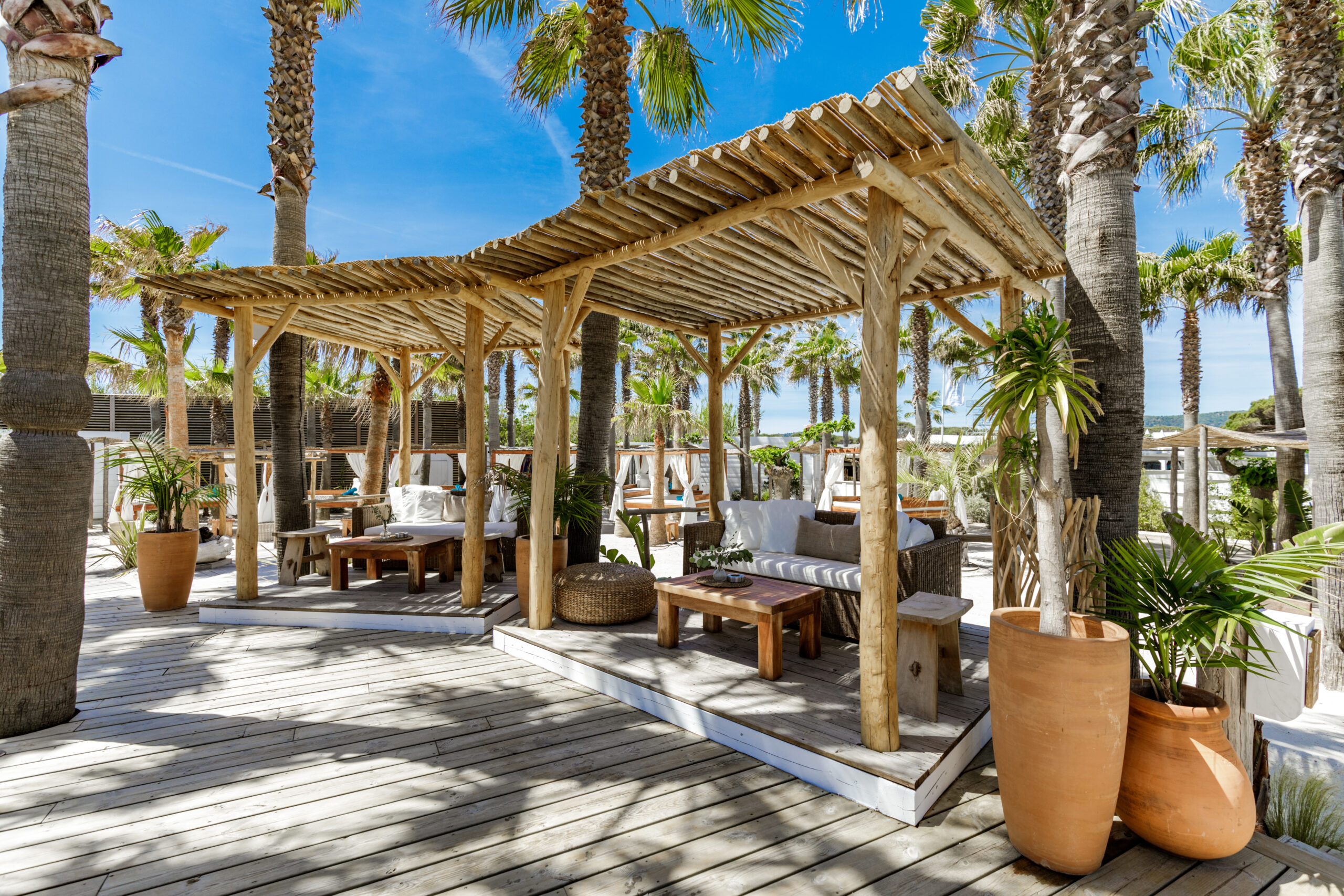 Des canapés, fauteuils et tables basses à l'ombre de treilles en bois et de palmiers, sur la plage privée de Nikki Beach, à St Tropez