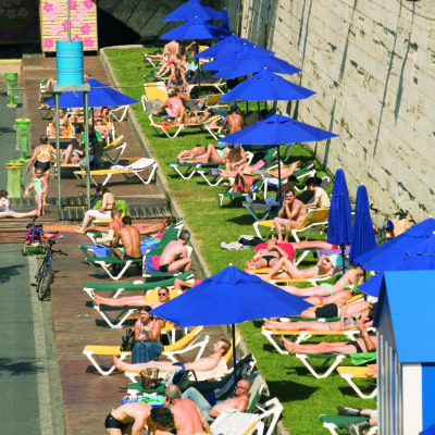 Un quai de la Seine à Paris, avec de nombreux parasols bleus et des transats jaunes sur lesquels des Parisiens sont couchés, en maillot de bain, lors des journée Paris Plage