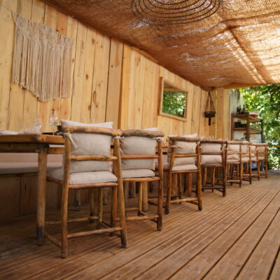 Des fauteuils en bois avec coussins en lin beige alignés face à des tables en bois, sous une sorte de treille tressée, dans le restaurant de la Beach Parisienne, la plage à Paris