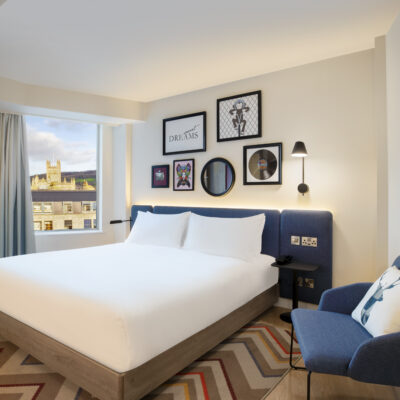 Photo d'une chambre d'un hôtel Hilton à Bath, avec un grand lit blanc, une tête de lit bleue assortie à un fauteuil bleu, des cadres au mur, et une fenêtre avec vue sur une église ou un château au beffroi carré