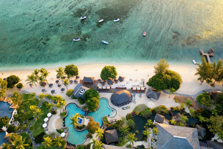 Vue aérienne d'un hôtel Hilton sur l'île Maurice avec plusieurs villas aux toits de palmes, une piscine aux formes arrondies entourée d'arbres, de palmiers, de transats et de parasols blancs. L'hôtel est au bord de l'océan Indien, le long d'une plage de sable blanc avec des transats