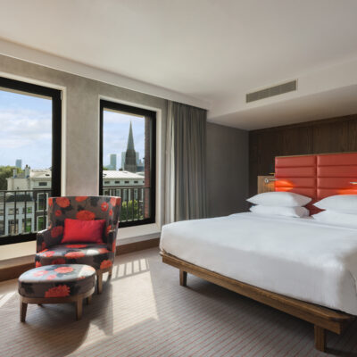 Photo d'une chambre d'un hôtel Hilton à La Hague, avec des murs gris, un grand lit blanc avec une tête de lit en cuir molletonné rouge assorti à un fauteuil gris avec des fleurs rouges, et deux fenêtres avec vue sur des immeubles blancs et un clocher