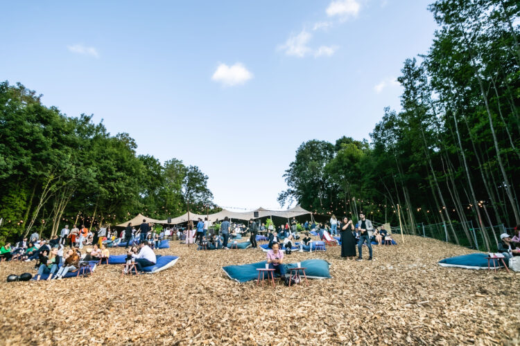 Une plage de copeaux de bois avec des jeunes assis sur des poufs bleus et en arrière plan, une grande tente du Perchoir Y entourée par les arbres de Meudon près de Paris