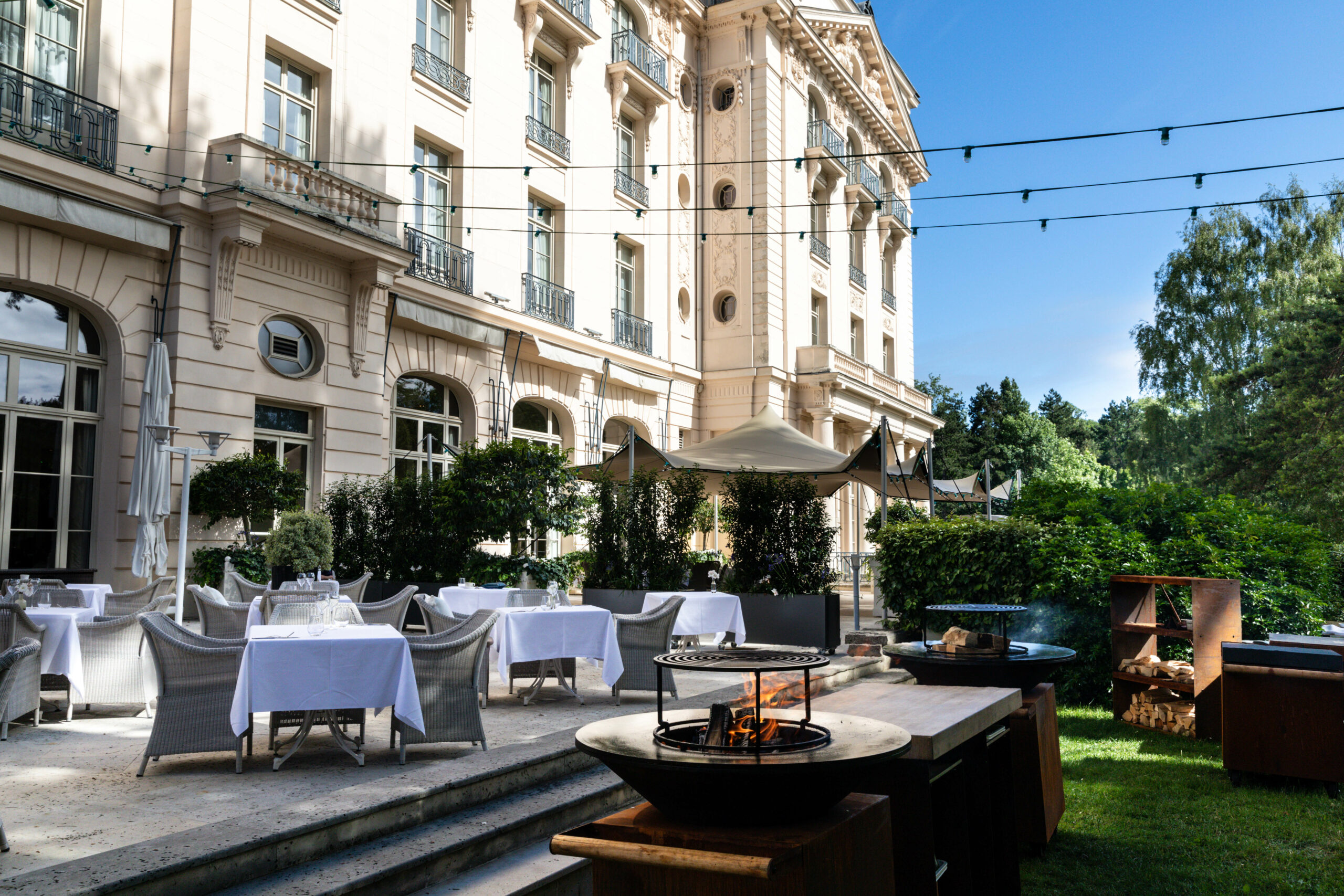 La façade éclairée du Trianon Palace Versailles avec en premier une terrasse où des tables sont élégamment dressées pour le diner, face à une plancha barbecue