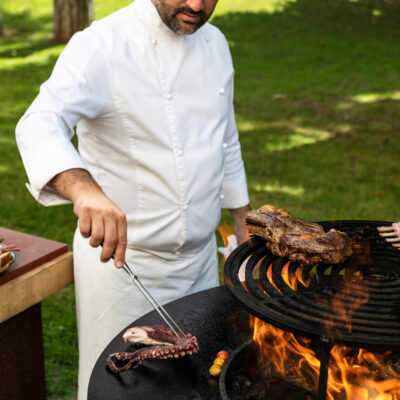 Un cuisinier du Trianon Palace Versailles en tenue de chef blanche, en train de faire griller des viandes sur une plancha ronde