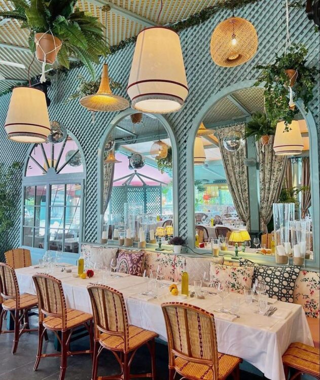 Vue intérieure du restaurant Le Sail Fish, avec ses grands miroirs aux murs, ses chaises tressées, sa longue table recouverte d'une nappe blanche, ses rideaux et coussins couverts de motifs fleuris