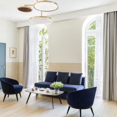 Une chambre de l'hôtel L'Imperator à Nîmes avec un canapé bleu et deux fauteuils assortis entourés par deux grandes fenêtres arrondies qui donnent sur les arbres d'un jardin