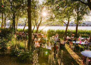 Une vue de la grande terrasse de Chez Alriqau bord de la Garonne, en face des quais de Bordeaux, avec sur le sable et sous les arbres, des tables et des chaises où sont installés de jeunes gens