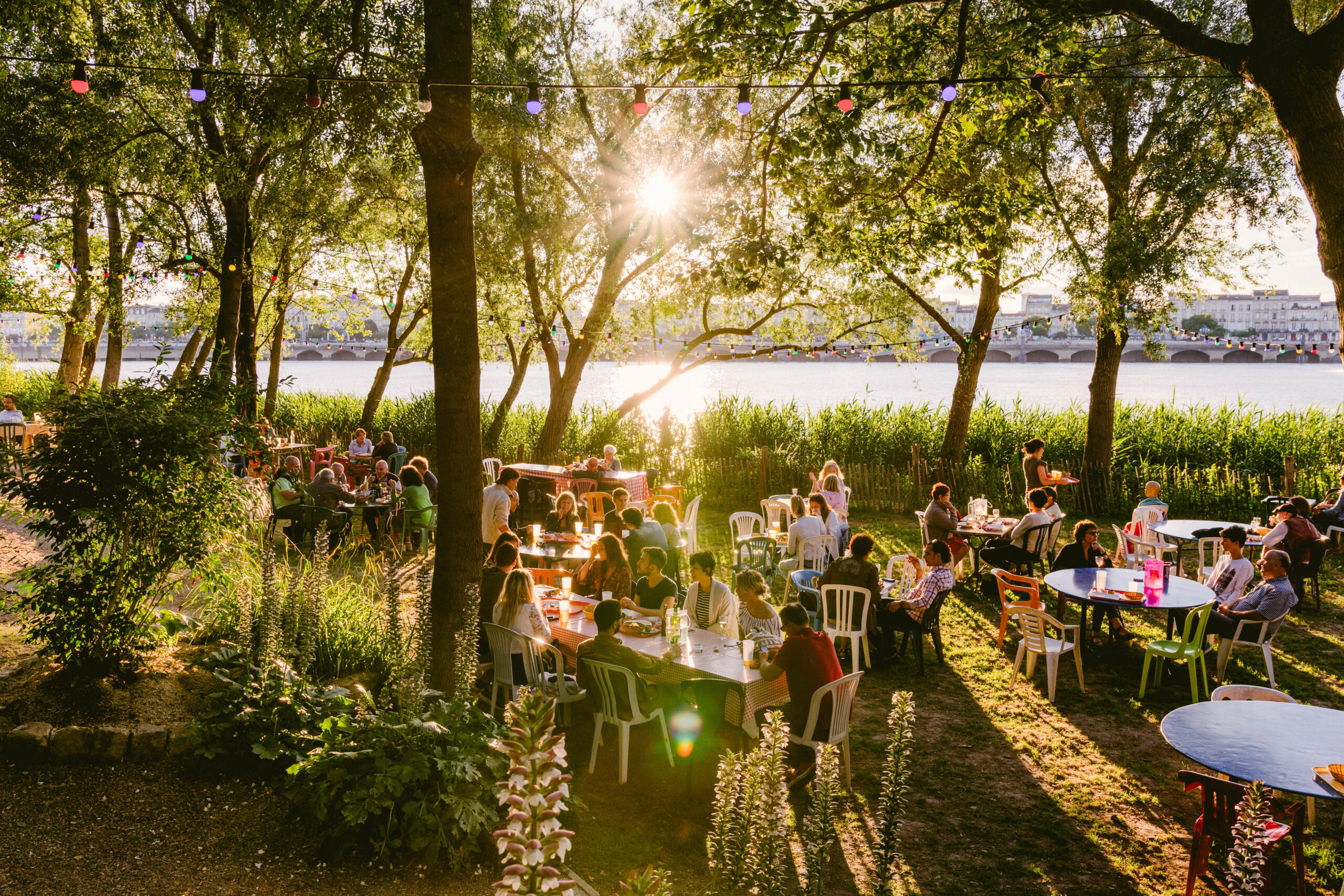 Une vue de la grande terrasse de Chez Alriqau bord de la Garonne, en face des quais de Bordeaux, avec sur le sable et sous les arbres, des tables et des chaises où sont installés de jeunes gens