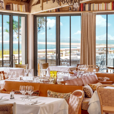 Le restaurant de La Co(o)rniche, avec ses fauteuils en cuir et ses tables élégamment dressées face à une vue sur le ciel bleu, la mer et le banc d'Arguin, dans le bassin d'Arcachon