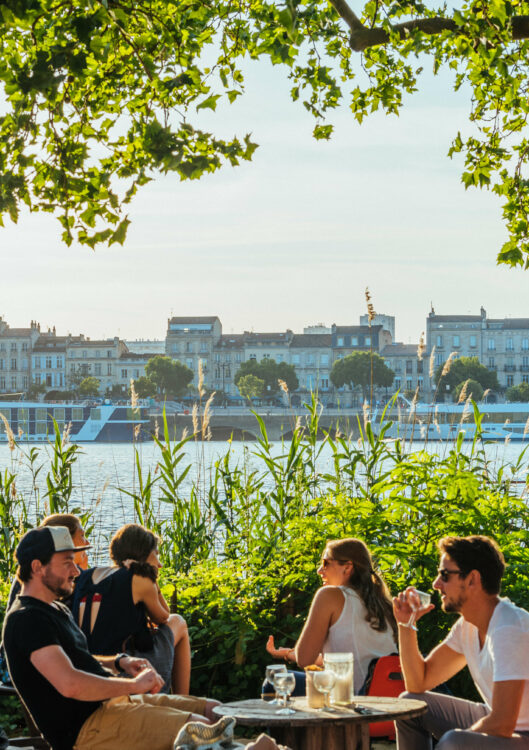 Des personnes attablées Chez Alriq, dans la verdure, au bord de la Garonne, avec au loin des quais du coeur historique de Bordeaux. 