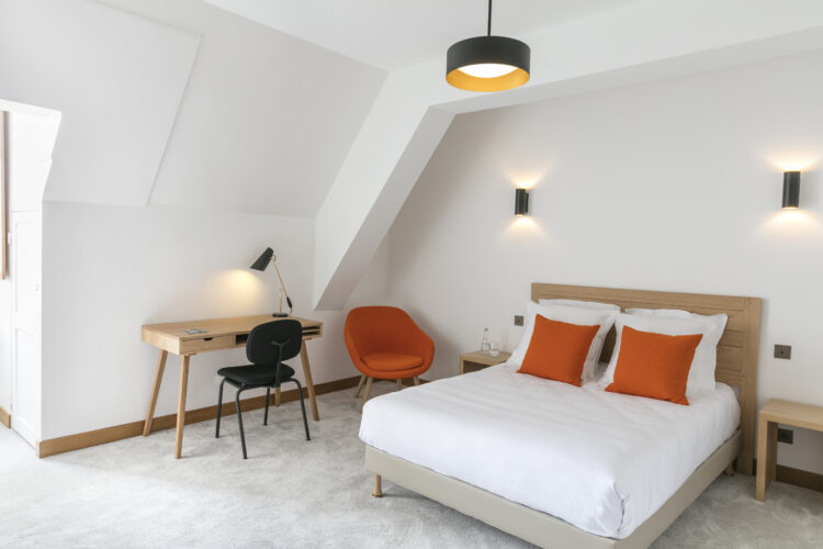 Une chambre de l'hôtellerie de Royaumont, toute blanche, avec un lit blanc, une chaise style années 50 en velours orange et un bureau en bois clair avec une chaise noire moderne, le tout éclairé par une petite fenêtre.