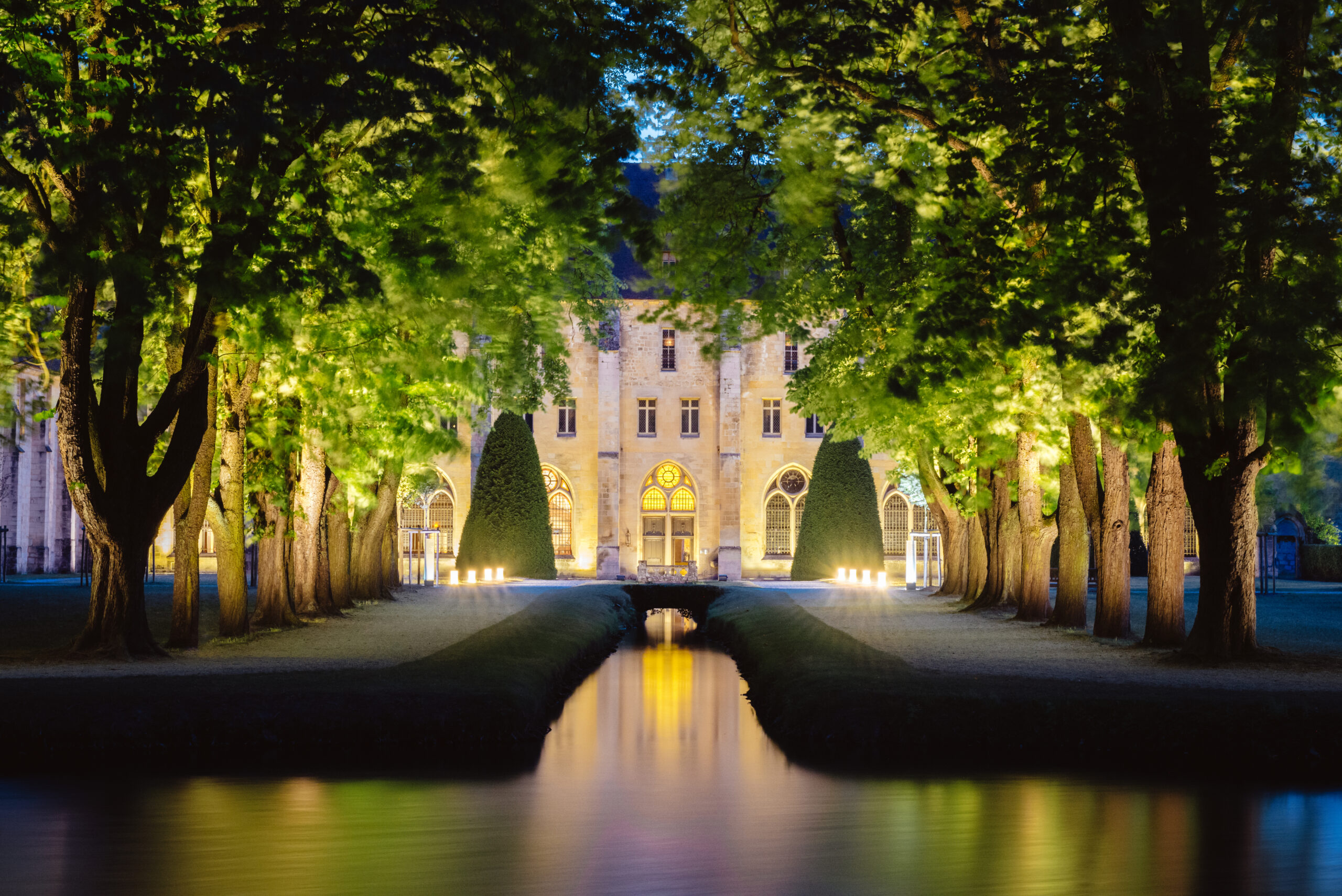 Une vue nocturne de l'abbaye de Royaumont, avec une allée qui entoure un bassin menant à la batisse