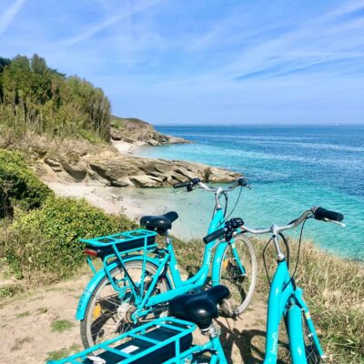Deux vélos électriques de location turquoise béquillés le long de la côte rocheuse, près d'une plage blonde de l'île de Groix