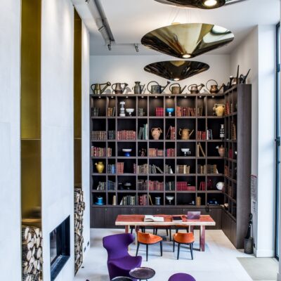 Vue du salon bibliothèque du Relais de Chambord, avec sa cheminée en insert contemporaine, sa haute bibliothèque chargée d'ouvrages, ces fauteuils violets et oranges, ces luminaires dorés futuristes