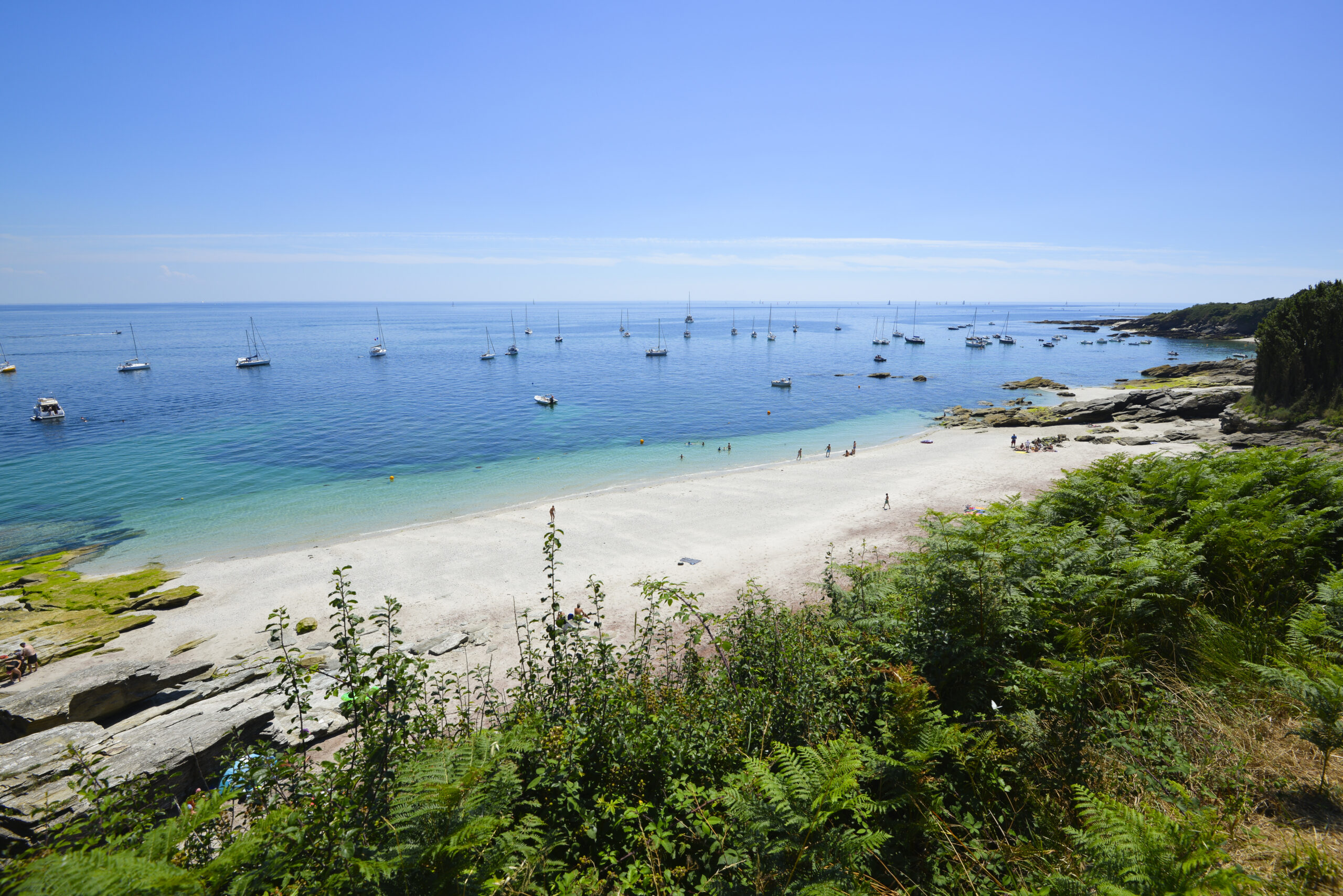 Vue, depuis le sentier de la plage Les Grands Sables sur l'île de Groix, avec son sable fin qui semble fondre dans la mer aux dégradés de bleus turquoise et indigo