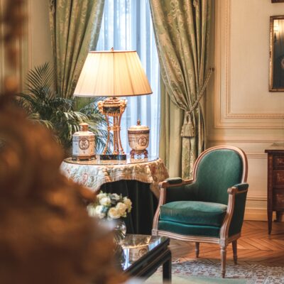 Détail d'une chambre du Relais & Château Domaine Les Crayères avec petite table ronde juponnée ornée de vases précieux, tapis persan et confortable fauteuil du 18e siècle en velours vert chasse