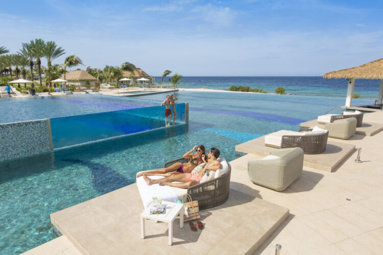 Un couple tendrement enlacé sur un sunbed, au bord d'une piscine qui déborde sur les cocotiers et une plage de sable blanc