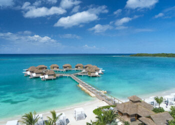 Une plage de sable blanc, des cocotiers, la mer des Caraïbes et le ciel bleu, un ponton qui mène aux villas sur pilotis de Sandals Resorts