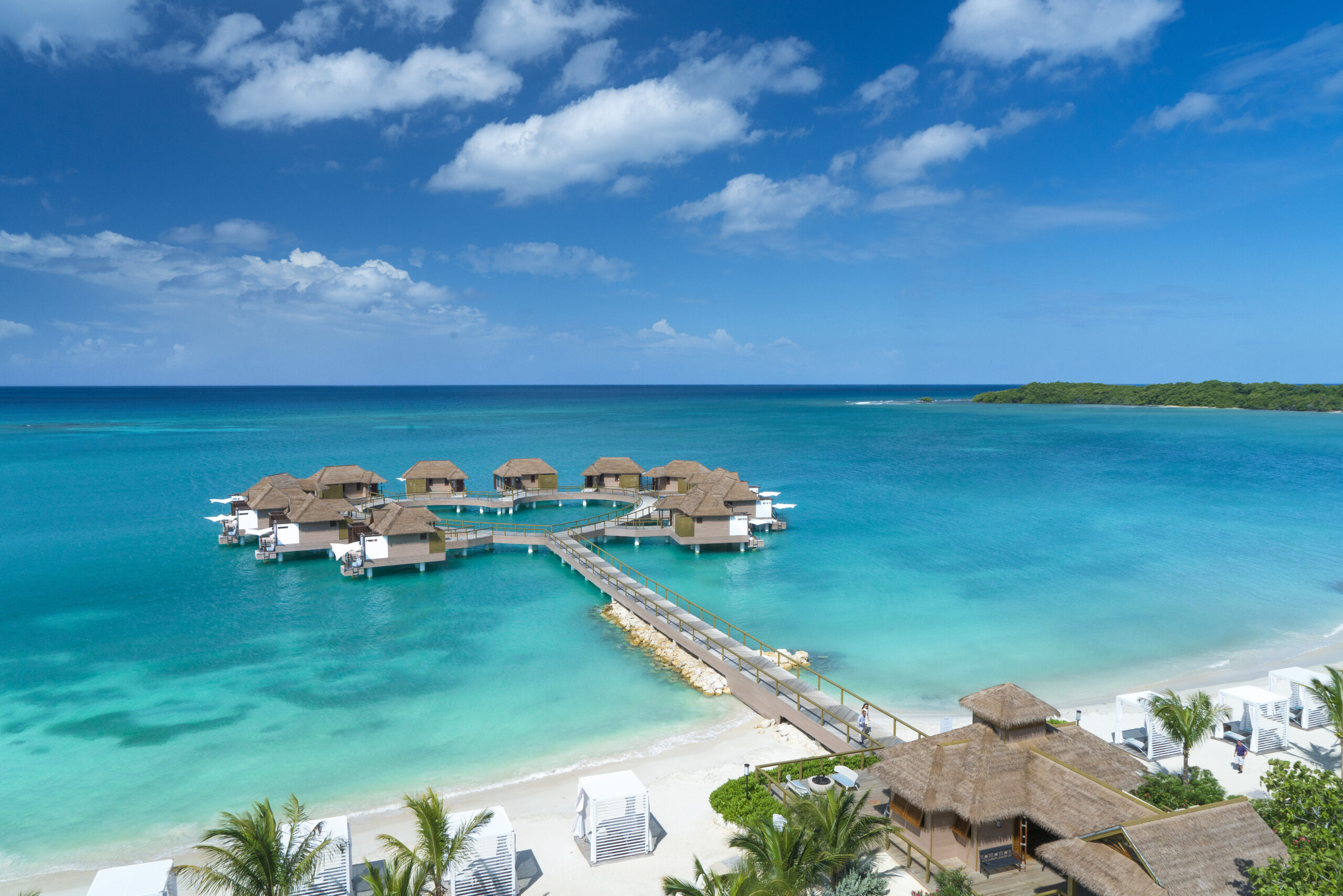 Une plage de sable blanc, des cocotiers, la mer des Caraïbes et le ciel bleu, un ponton qui mène aux villas sur pilotis de Sandals Resorts